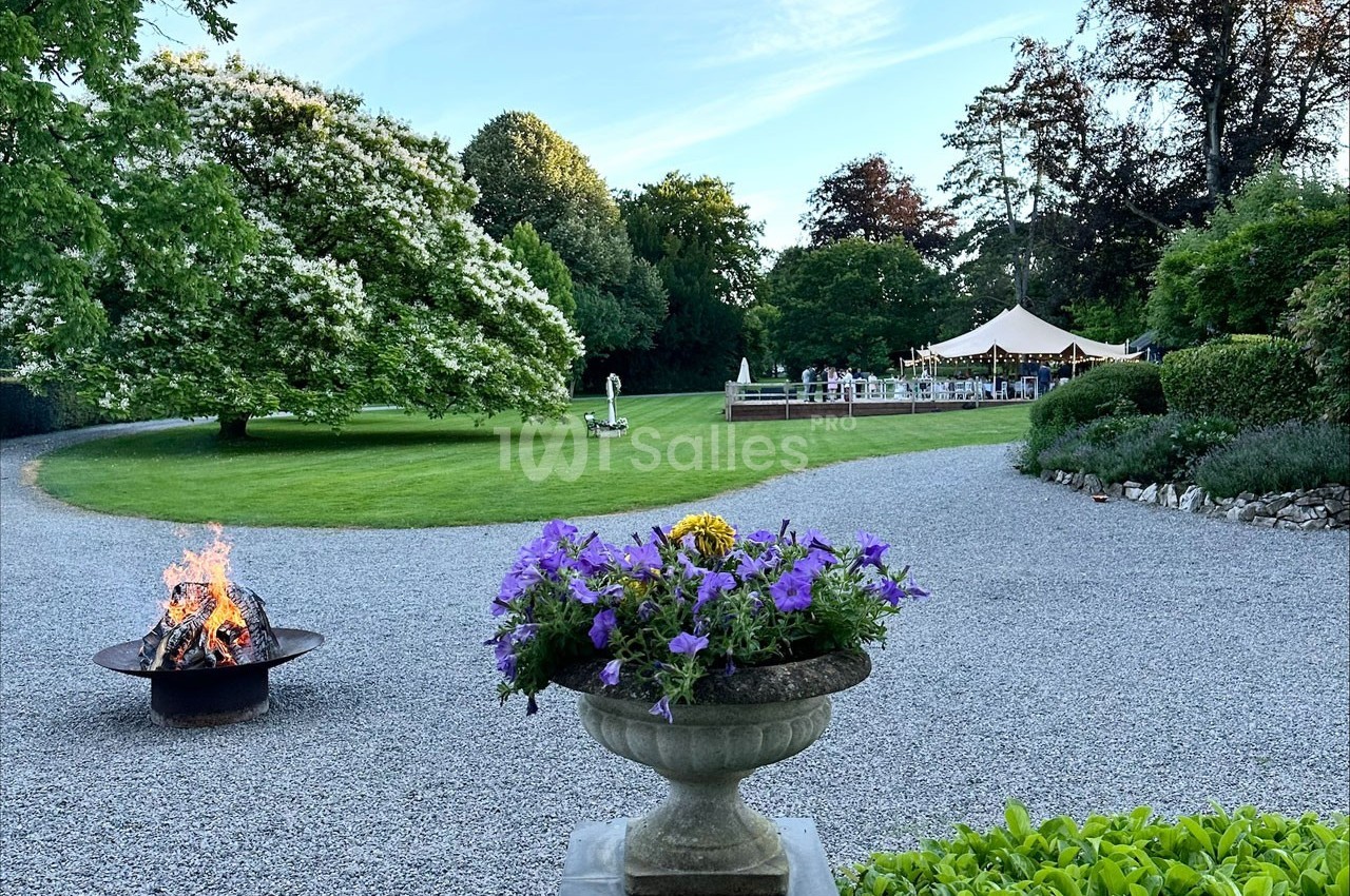 Allée gravillonnée bordée de verdure, avec un brasero, une jardinière fleurie et une tente au fond dans un parc.