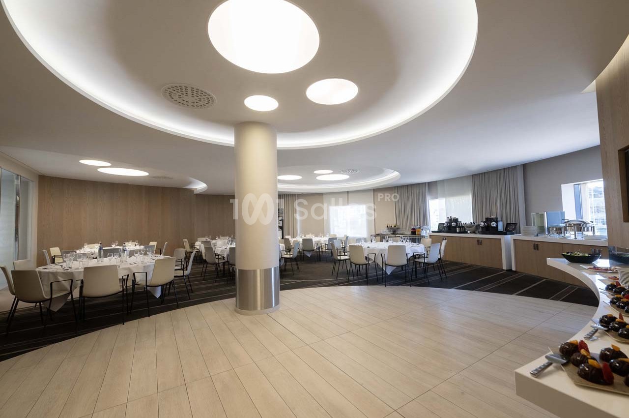 Salle de réception lumineuse avec tables dressées, buffet en arrière-plan et éclairage moderne au plafond.