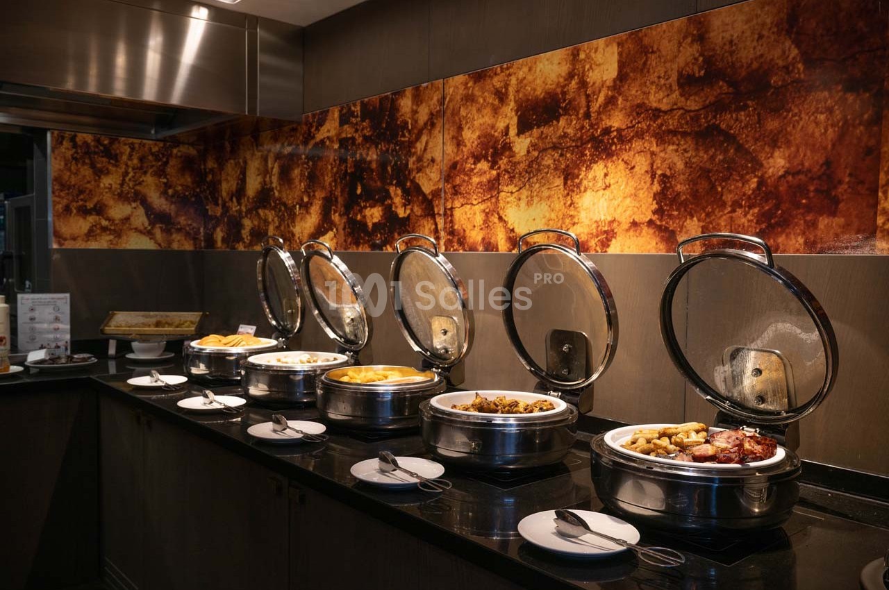 Buffet chaud avec plusieurs plats disposés dans des récipients en inox sur un comptoir sombre, décor mural texturé.
