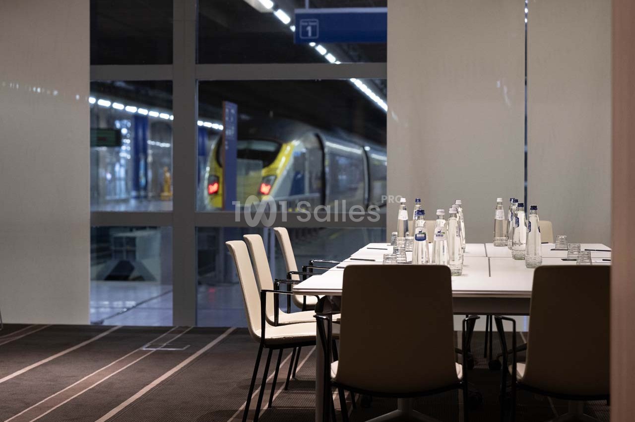 Salle de réunion moderne avec table et chaises, vue sur un train à quai derrière une baie vitrée.