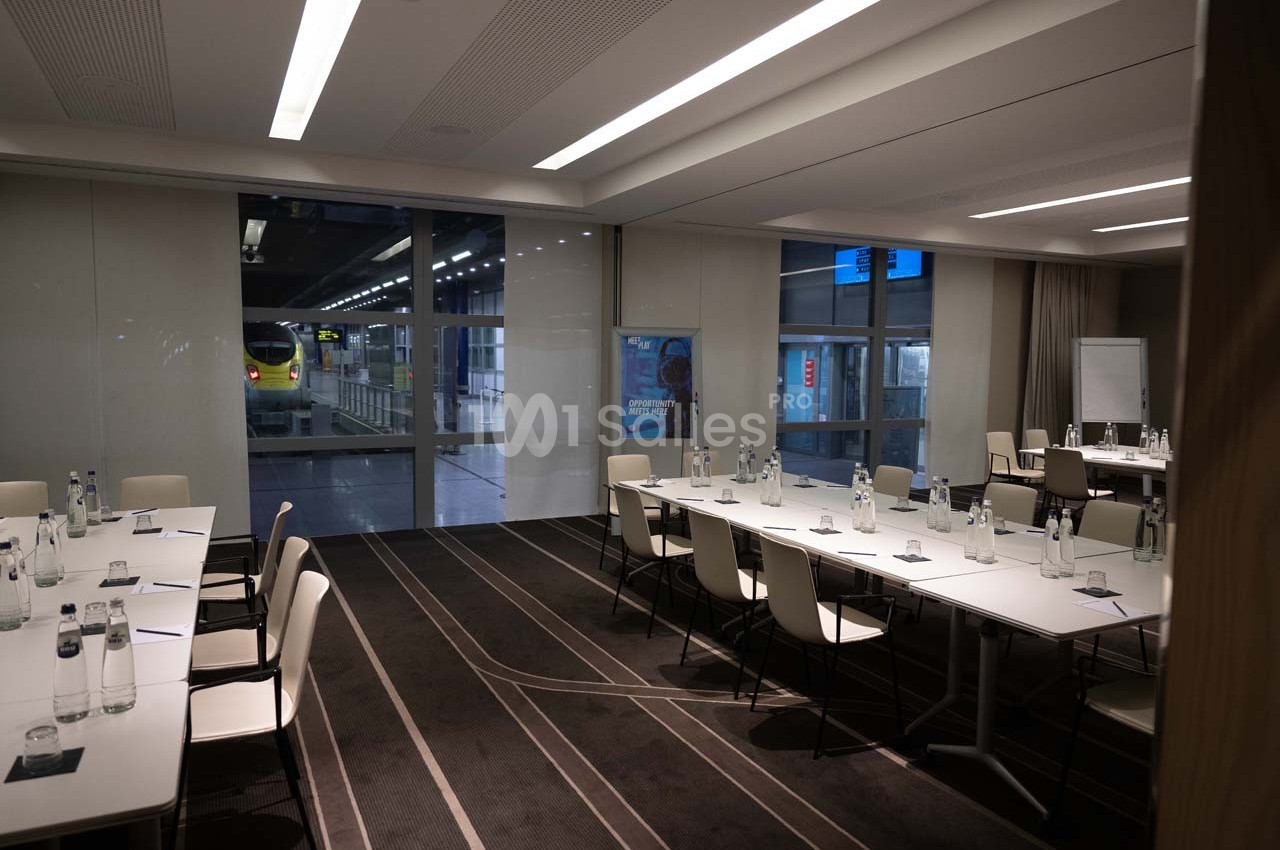 Salle de réunion moderne avec tables disposées en U, chaises blanches et vue sur une gare à travers de grandes baies vitrées.
