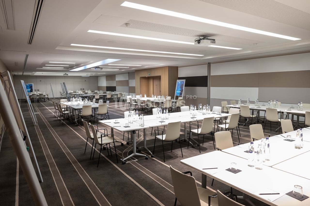 Salle de réunion moderne avec tables disposées en U, chaises alignées, bouteilles d'eau et écrans de présentation.