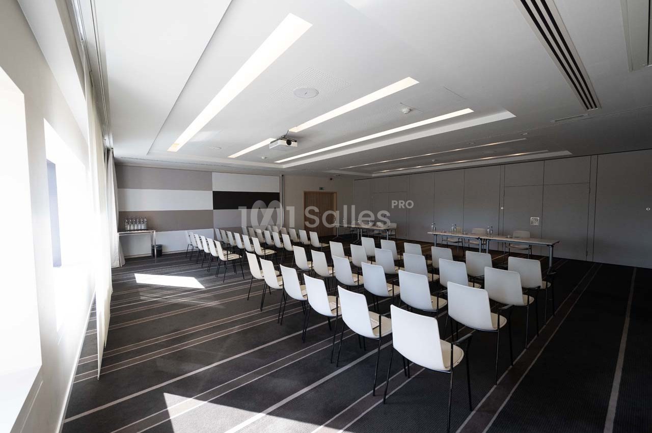 Salle de conférence lumineuse avec des rangées de chaises blanches alignées face à une table et un écran.