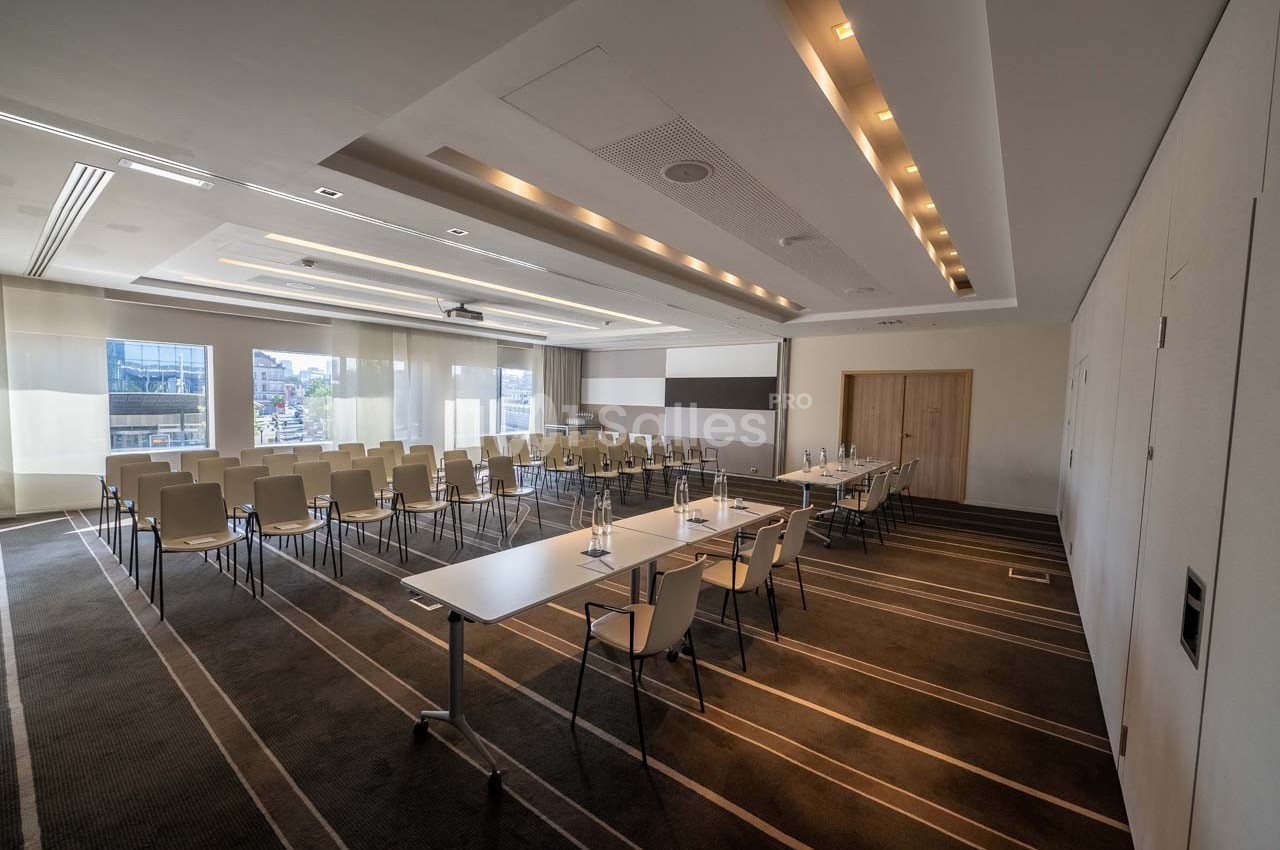 Salle de conférence lumineuse avec chaises alignées, tables équipées de verres et stylos, et grandes fenêtres vitrées.