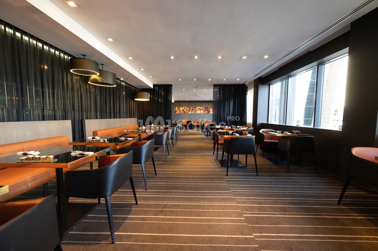 Salle de restaurant moderne avec tables dressées, chaises en cuir et grandes baies vitrées laissant entrer la lumière…