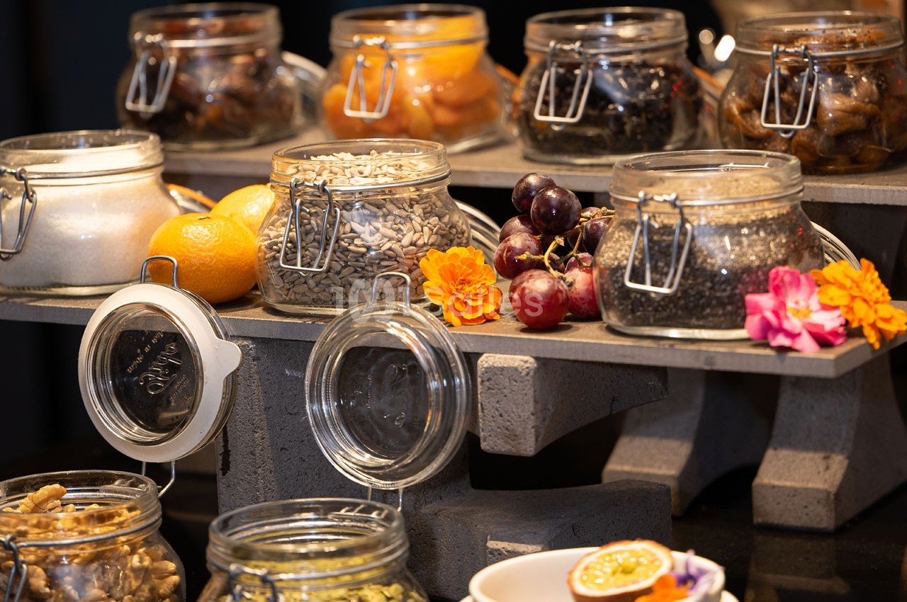 Bocaux en verre contenant graines, fruits secs et confits, disposés sur un présentoir en pierre avec décorations florales.