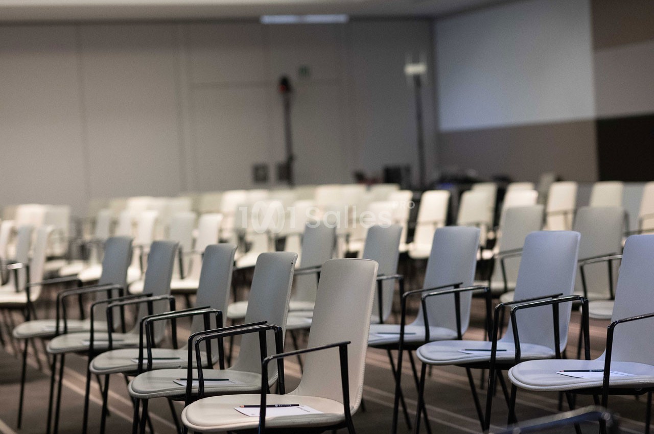 Salle de conférence avec des rangées de chaises blanches et noires alignées, certaines avec des documents posés dessus.