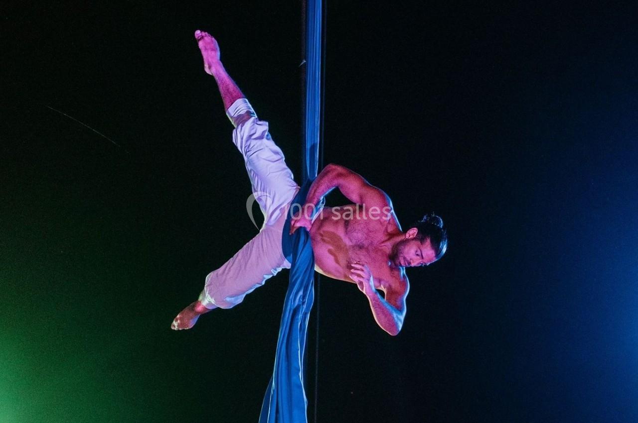 Un acrobate torse nu réalise une figure suspendu à un tissu aérien dans un environnement sombre éclairé par des lumières…