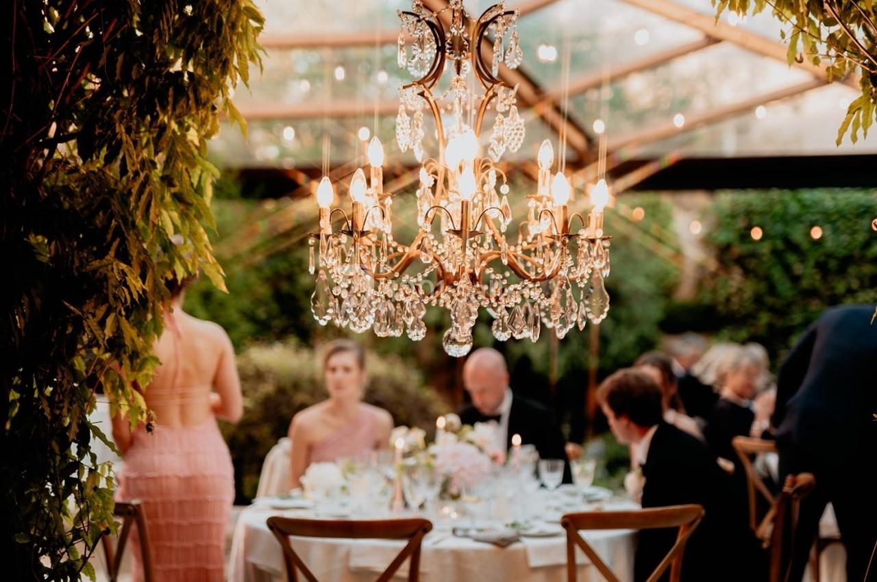 Lustre en cristal allumé suspendu sous une verrière, éclairant des convives assis à une table dans un jardin.