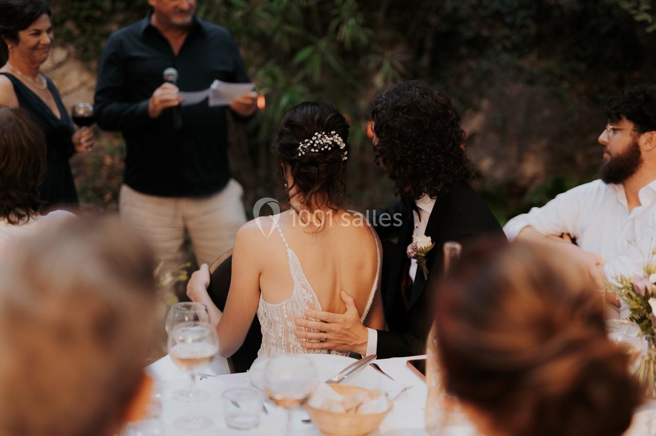 Un couple assis à une table lors d'un discours, entouré d'invités dans un cadre extérieur.