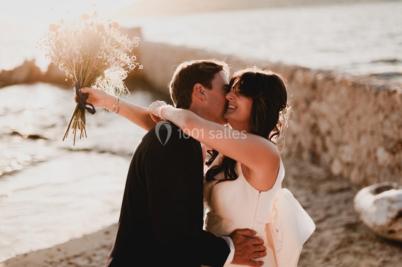 Un couple en tenue de mariage s'embrasse sur une plage au coucher du soleil, la mariée tenant un bouquet.