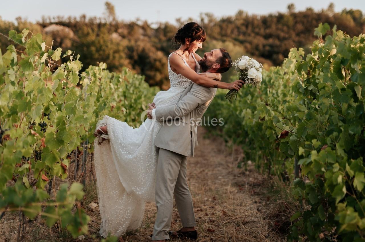 Un couple en tenue de mariage pose dans un vignoble, l'homme portant la femme dans ses bras.
