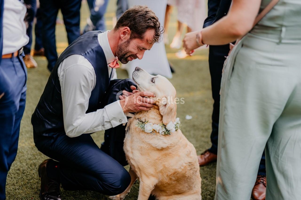Un homme en costume caresse un labrador portant un collier de fleurs lors d'un événement en extérieur.