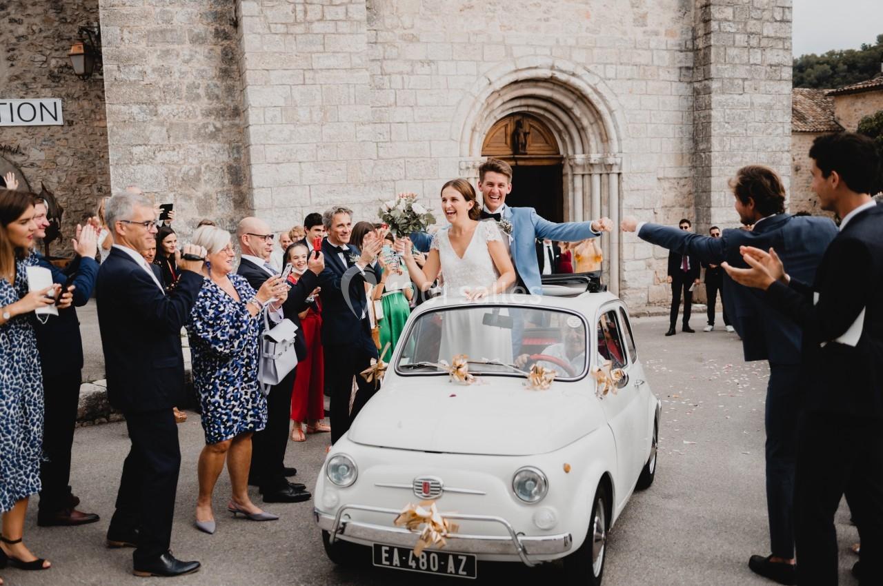 Un couple de mariés souriants quitte une cérémonie dans une petite voiture blanche, entouré d'invités applaudissant.