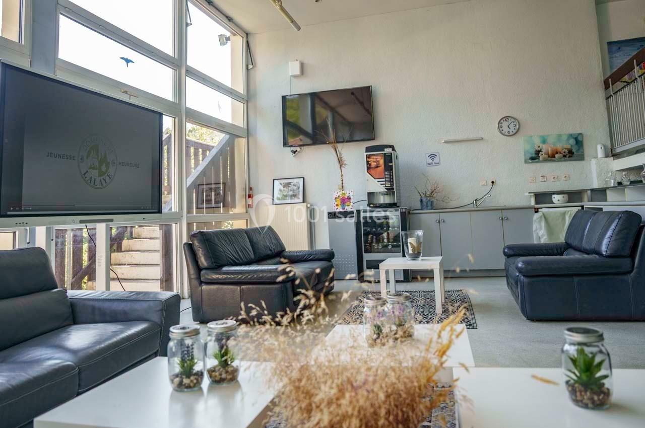 Salle de détente lumineuse avec canapés, écran, machine à café et décoration végétale sur une table basse.
