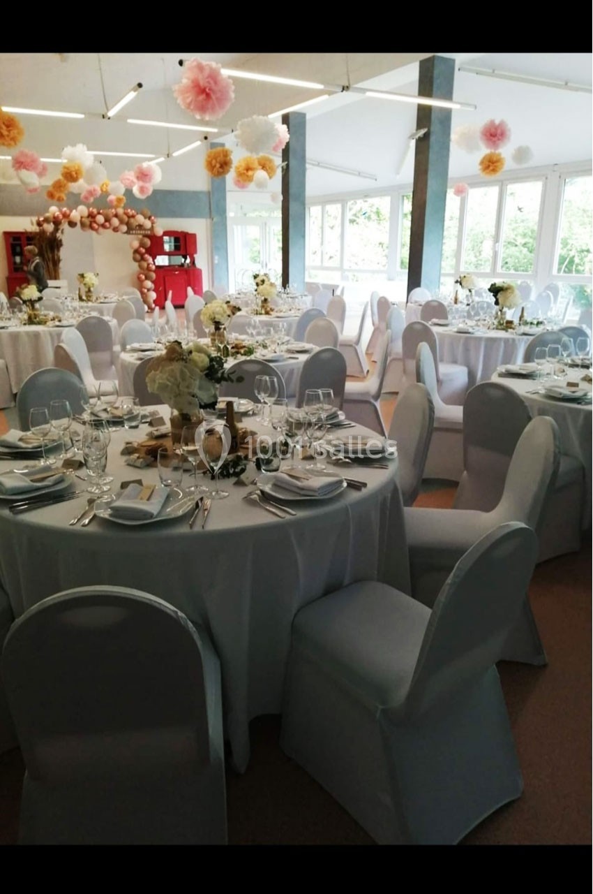 Salle de réception décorée avec des tables rondes dressées, des chaises blanches et des guirlandes de fleurs suspendues.