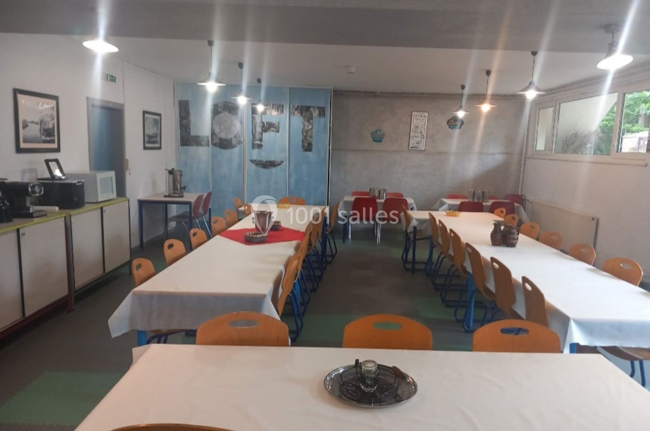 Salle à manger lumineuse avec de longues tables recouvertes de nappes blanches, entourées de chaises colorées.