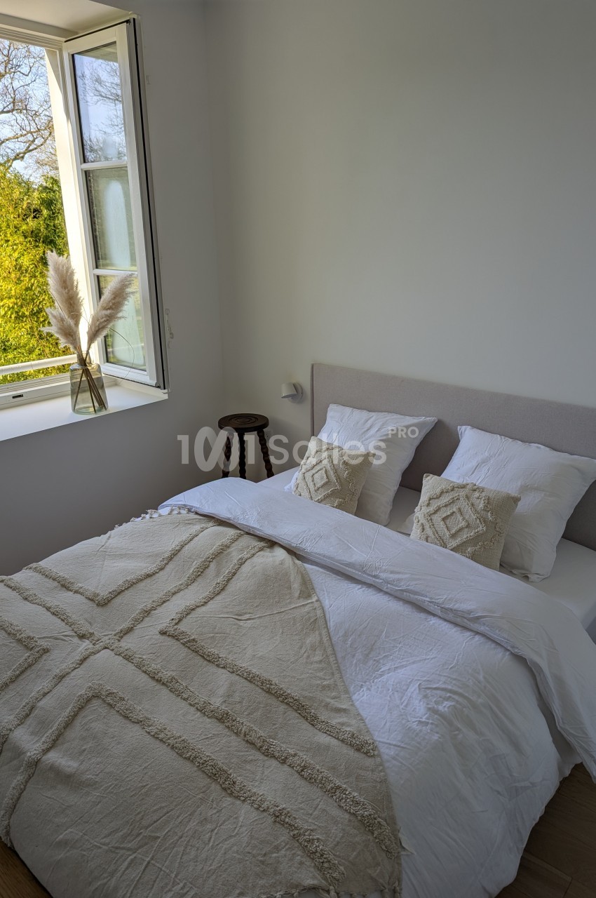 Lit double avec draps blancs et plaid beige, près d'une fenêtre ouverte donnant sur un jardin verdoyant.