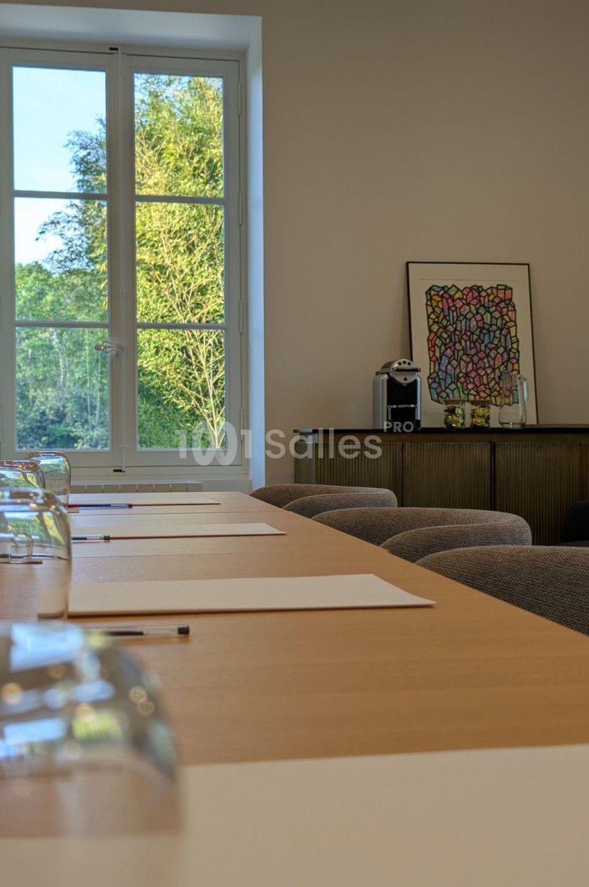 Salle lumineuse avec une table en bois, des feuilles et crayons, vue sur un jardin par une grande fenêtre.