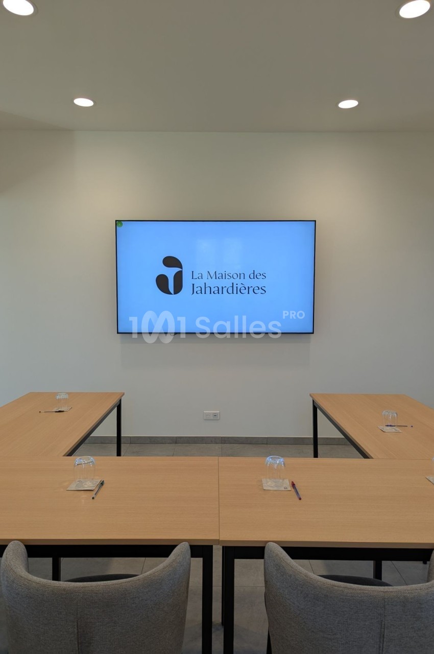 Salle de réunion lumineuse avec tables en U, chaises grises et écran affichant ’La Maison des Jahardières’.