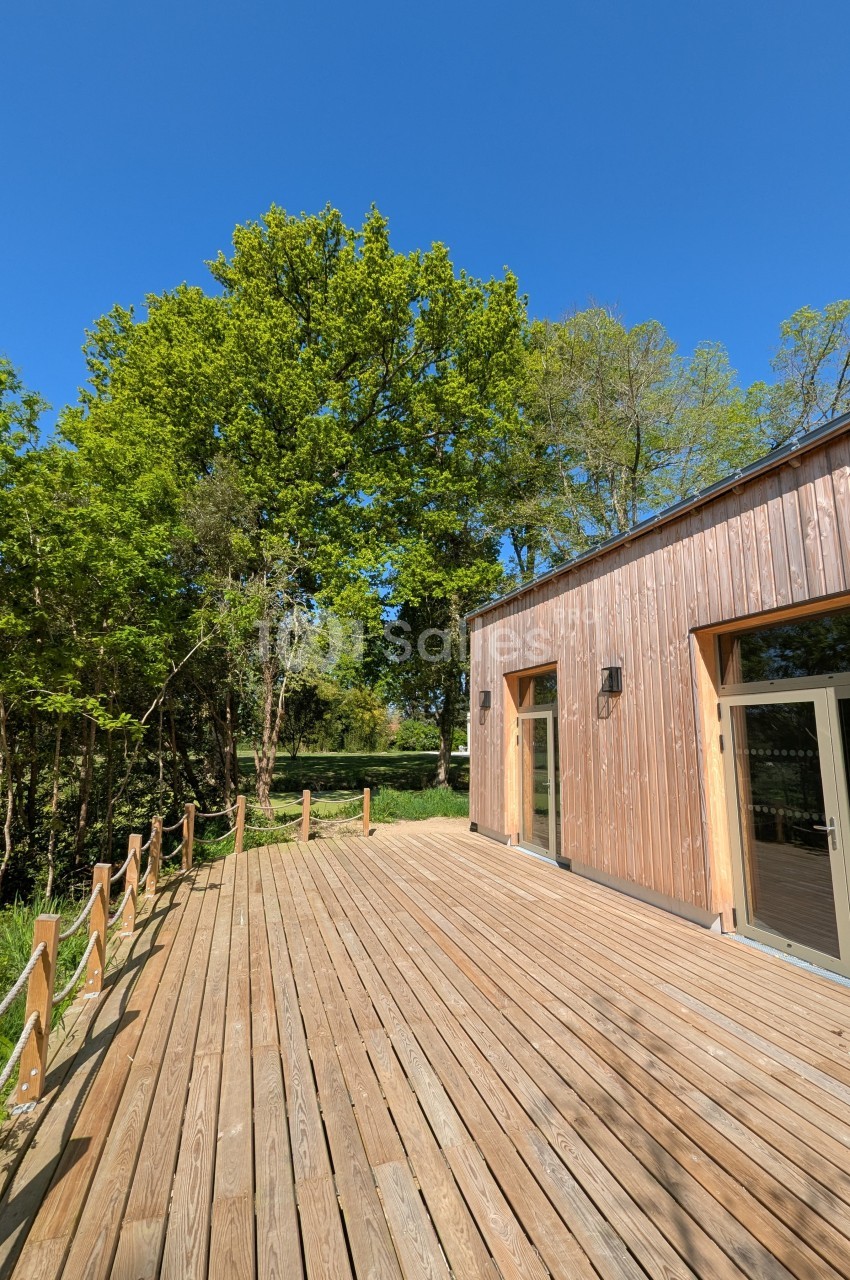 Terrasse en bois bordée de cordes, jouxtant un bâtiment en bois, entourée d'arbres sous un ciel bleu.