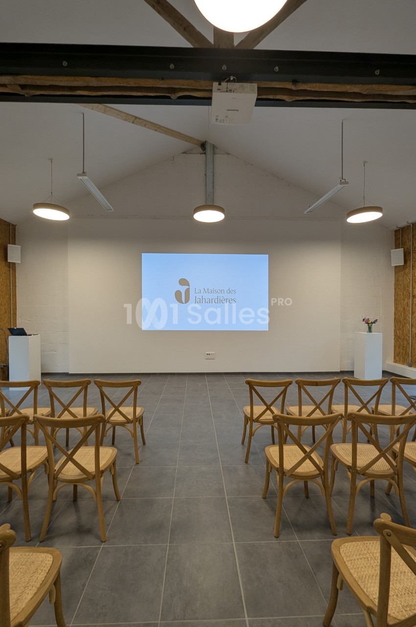 Salle de conférence avec écran de projection allumé, rangées de chaises en bois et éclairage suspendu.