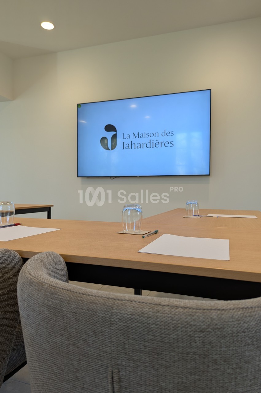Salle de réunion avec écran affichant le logo ’La Maison des Jahardières’, tables équipées de papiers, stylos et verres.