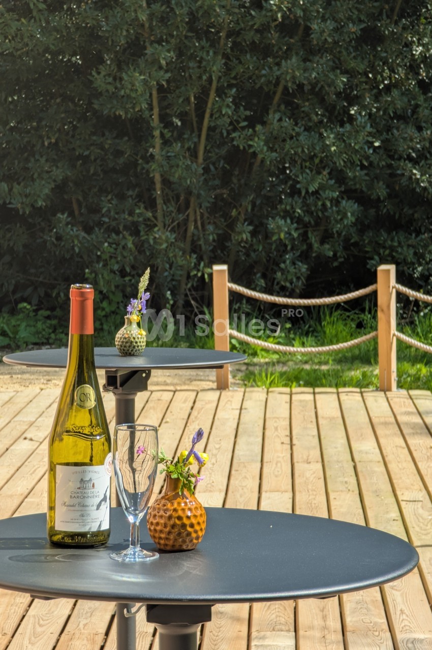 Bouteille de vin, verre et petites décorations florales sur une table ronde en extérieur, avec terrasse en bois et…