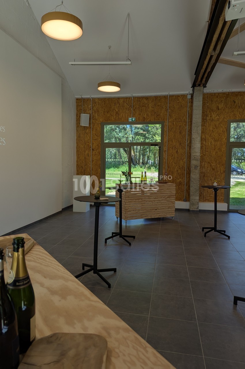 Salle lumineuse avec tables hautes, bouteilles de champagne et vue sur un espace vert à travers de grandes fenêtres.