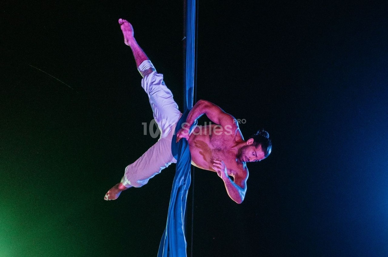 Un homme torse nu réalise une figure acrobatique suspendu à un tissu aérien, sur fond sombre.