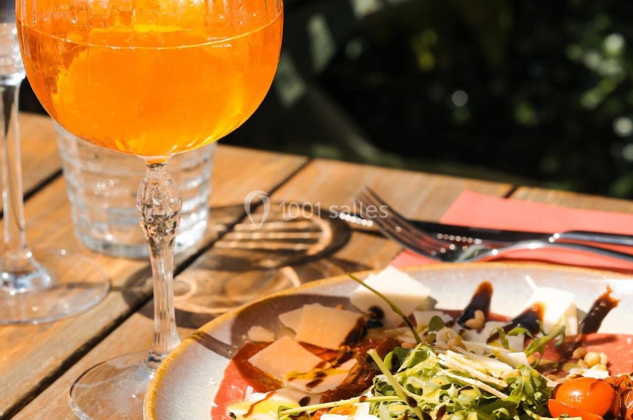 Un verre de cocktail orange et une assiette de carpaccio avec roquette et copeaux de fromage sur une table en bois.