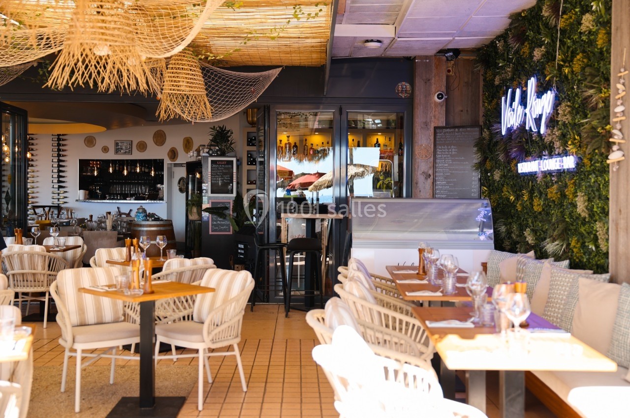 Salle de restaurant lumineuse avec tables dressées, décoration végétale et vue sur un bar en arrière-plan.