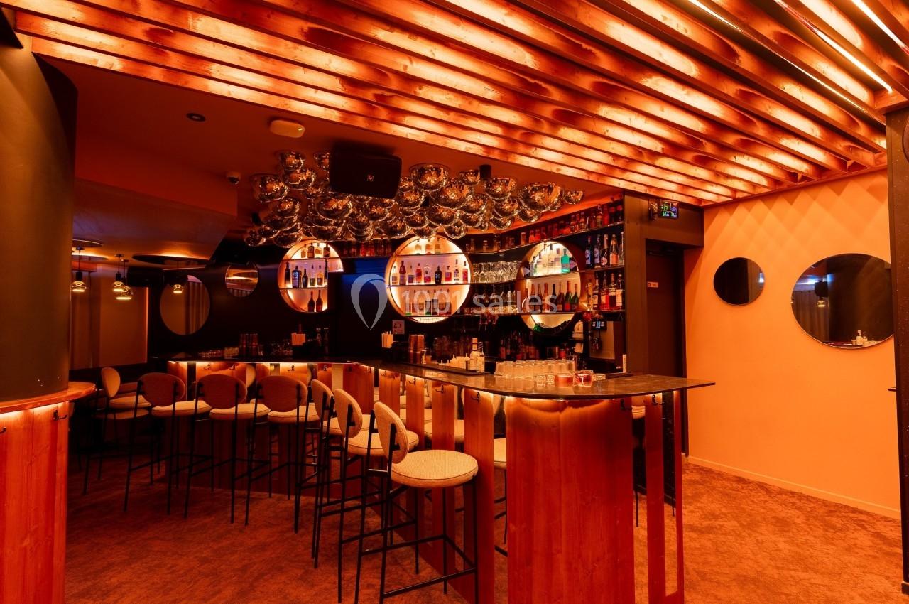 Bar moderne avec éclairage chaleureux, tabourets hauts, comptoir en bois et étagères remplies de bouteilles.