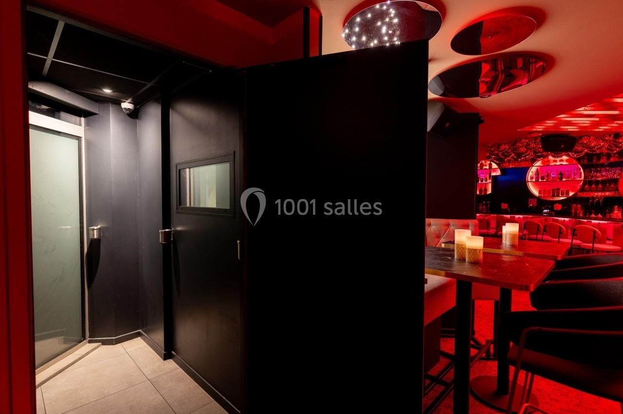 Entrée d'une pièce sombre avec une porte noire, à côté d'un espace de bar éclairé par des lumières rouges.