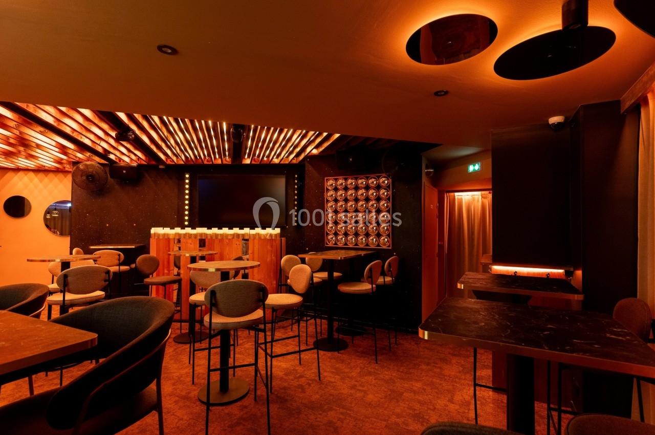 Salle de bar moderne avec éclairage chaleureux, tables hautes, chaises rembourrées et écran au fond.