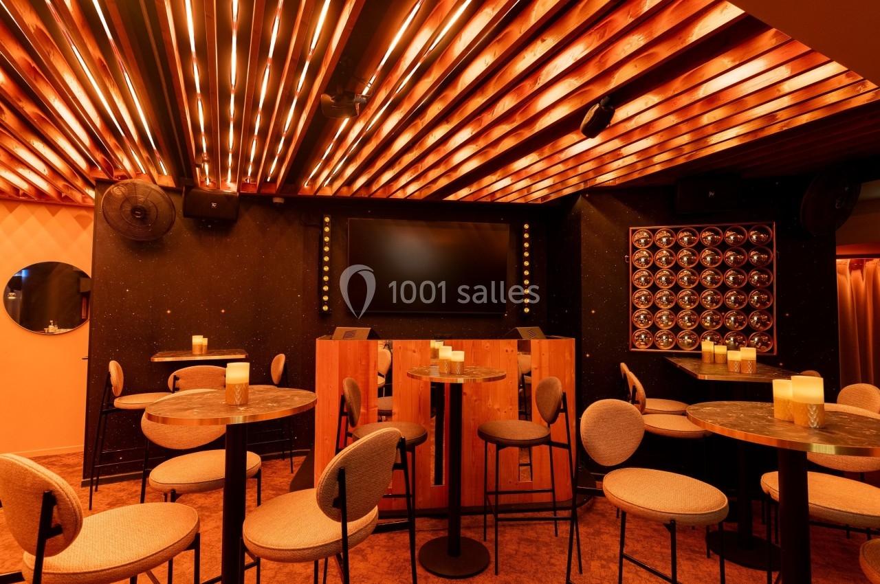 Salle de bar avec tables hautes, chaises rembourrées, éclairage chaleureux et écran mural noir.