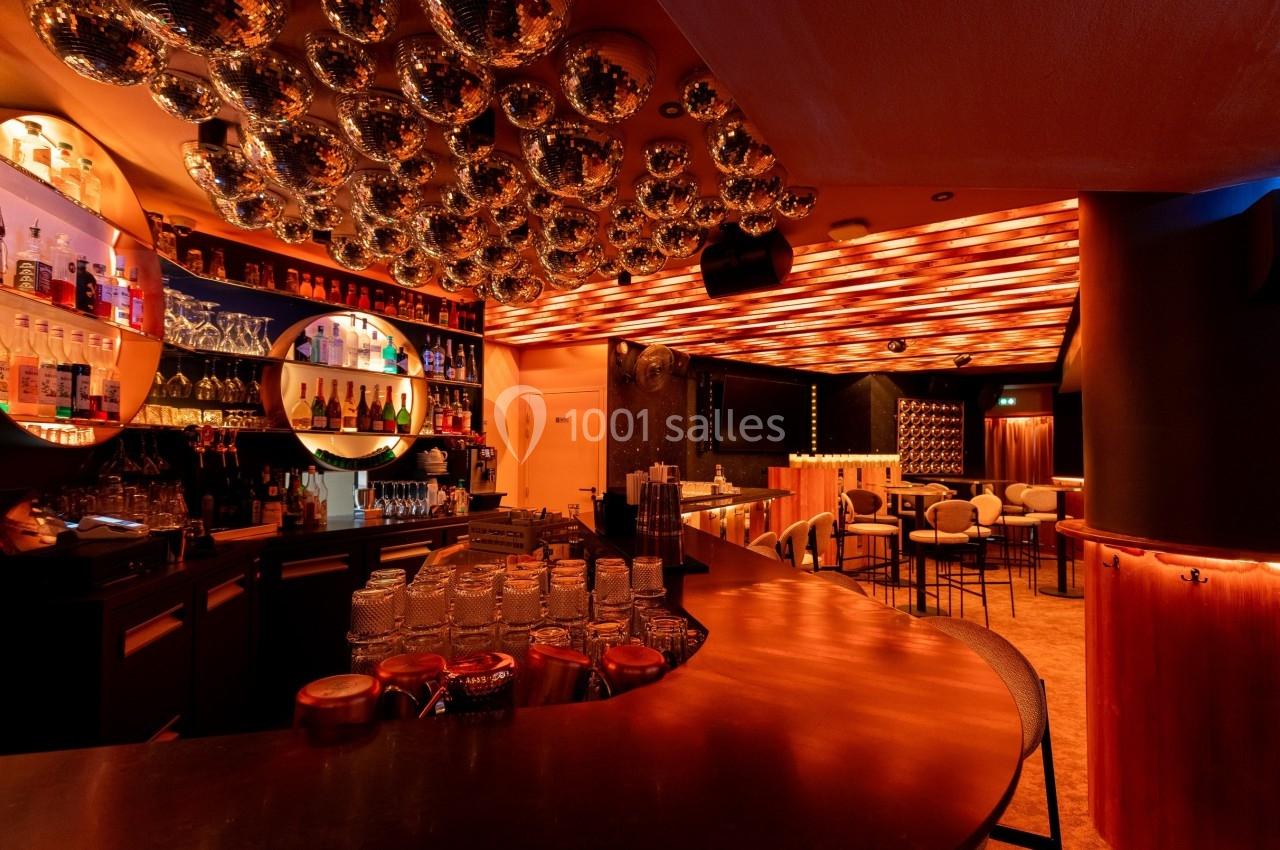 Intérieur d'un bar avec comptoir en cuivre, étagères de bouteilles illuminées et plafond décoré de sphères réfléchissantes.