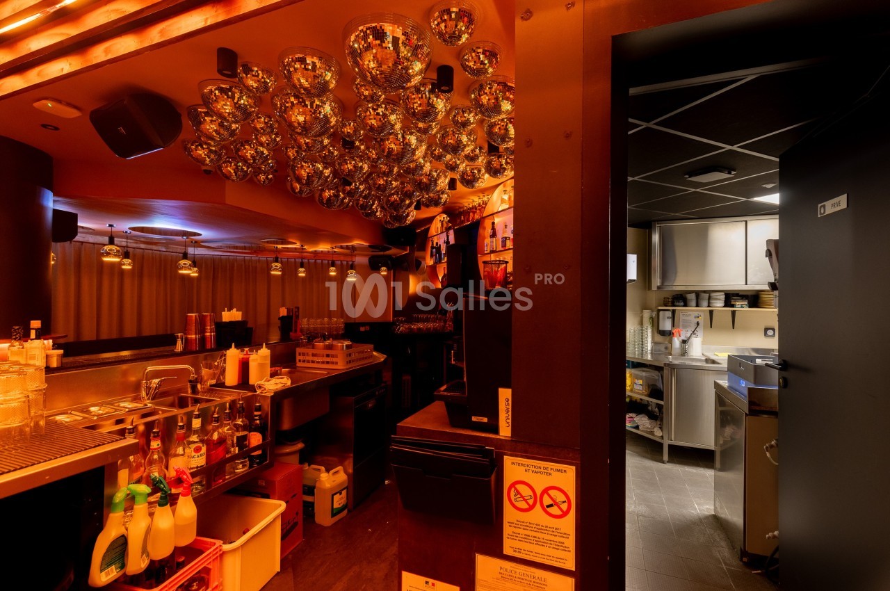Intérieur d'un bar avec comptoir équipé, éclairage chaleureux et accès visible à une cuisine professionnelle.