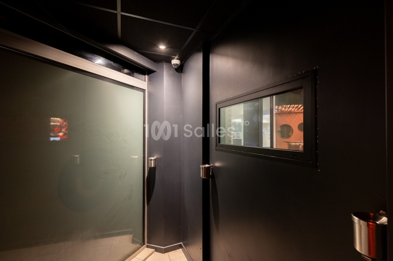 Vue d'un couloir sombre avec une porte vitrée givrée à gauche et une fenêtre rectangulaire sur un mur noir à droite.