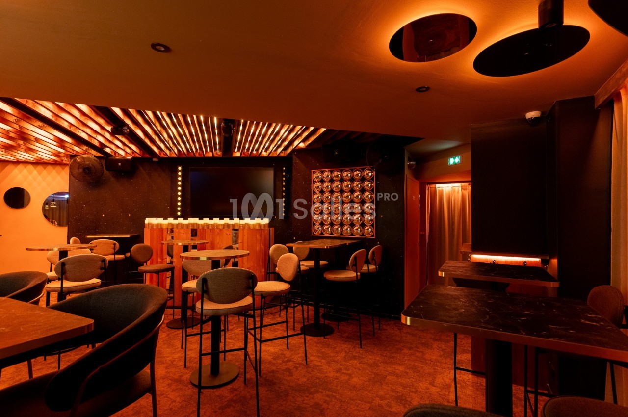 Salle de bar moderne avec éclairage chaleureux, tables hautes, chaises rembourrées et écran mural au fond.