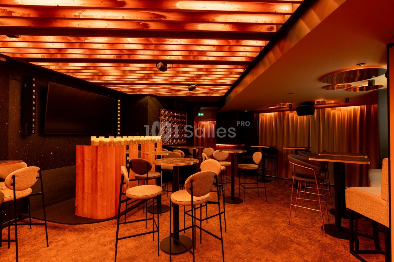Salle de bar avec éclairage chaleureux, tables hautes, chaises rembourrées et plafond en bois illuminé.