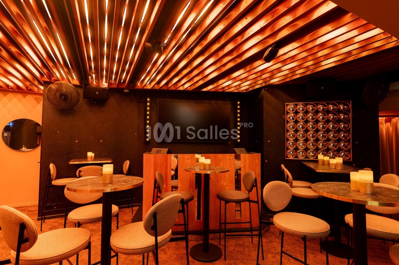Salle avec tables hautes, chaises rembourrées, éclairage chaleureux au plafond et écran noir sur un mur sombre.