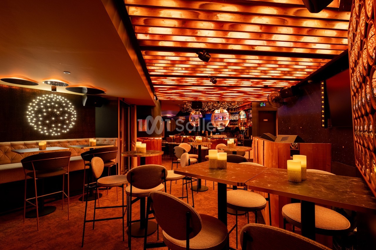 Salle de bar avec éclairage chaleureux, tables hautes, chaises rembourrées et décorations lumineuses au plafond.