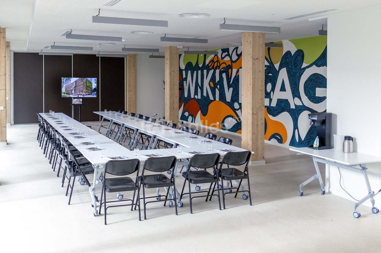 Salle de réunion lumineuse avec longues tables, chaises noires, mur décoré de graffitis et écran de présentation.