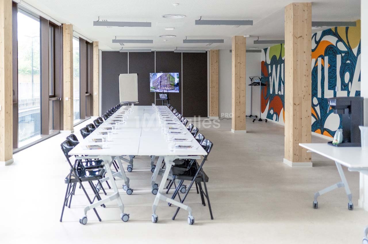 Salle de réunion lumineuse avec une longue table, des chaises noires, un écran, un tableau blanc et une fresque murale…