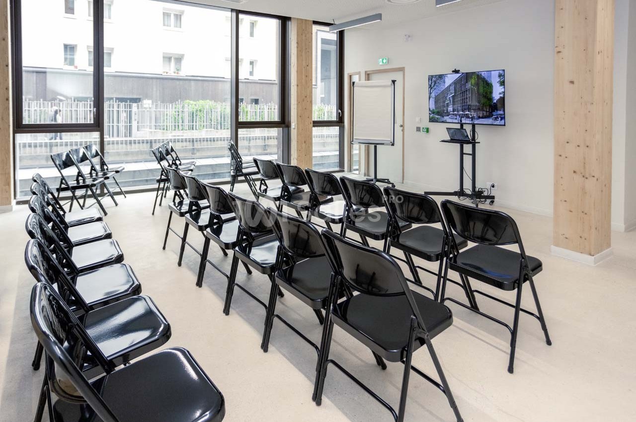 Salle de réunion lumineuse avec des chaises pliantes noires disposées en rangées face à un écran de présentation.