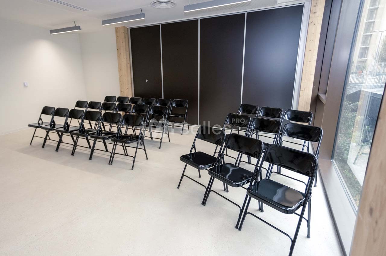 Salle lumineuse avec rangées de chaises pliantes noires alignées face à un mur sombre et une grande fenêtre.