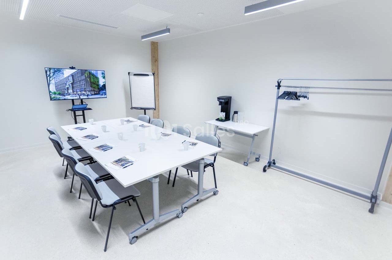 Salle de réunion lumineuse avec table, chaises, écran, paperboard, porte-manteaux et machine à café.