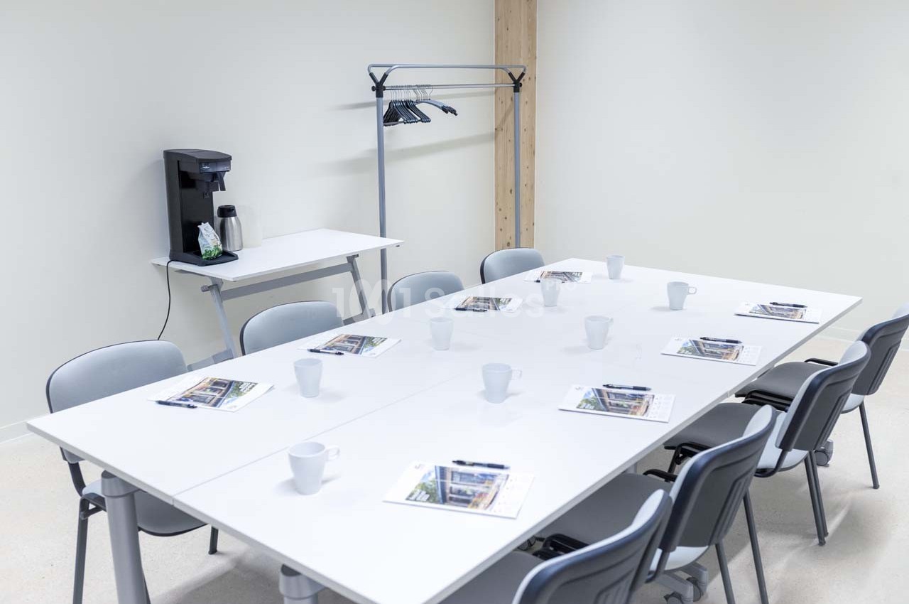 Salle de réunion avec table blanche, chaises grises, documents, tasses et une machine à café en arrière-plan.