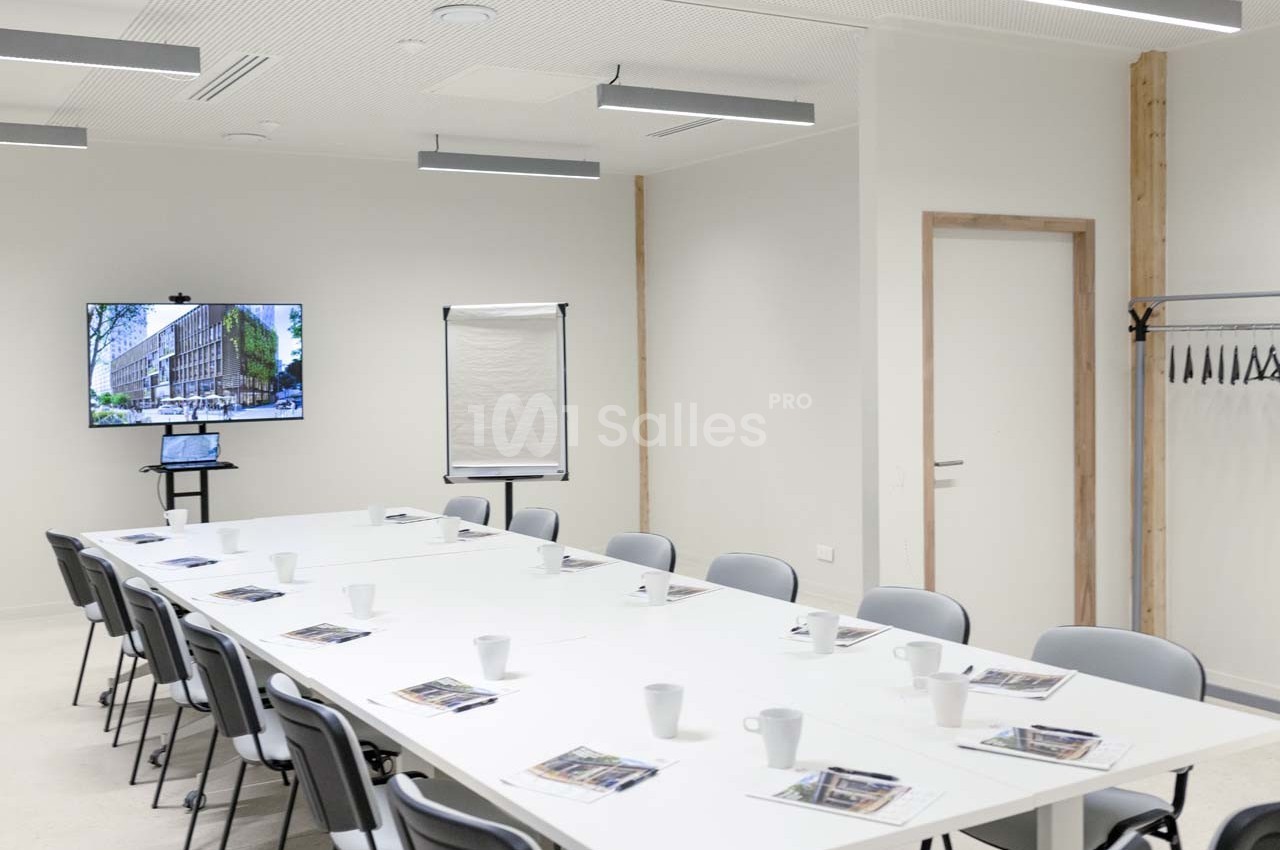 Salle de réunion lumineuse avec table en U, chaises, écran affichant un bâtiment, paperboard et porte-manteaux.